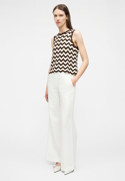 Γυναίκα που φοράει καφέ και λευκή αμάνικη μπλούζα με ζιγκ-ζαγκ σχέδιο και λευκό παντελόνι υψηλής μέσης με φαρδιά πόδια, συνοδευόμενο από λευκά μυτερά παπούτσια.