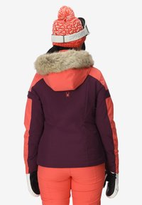 Giacca da sci viola con dettagli corallo e cappuccio in pelliccia sintetica. Indossata con pantaloni da sci arancioni e guanti bianchi, con un berretto di maglia con logo.