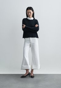 Juodas megztinis su raštuotu apačia, derinamas su baltais plačiomis kojomis džinsais, kurių rankogaliai sulankstyti, ir juodais aukštakulniais su smailiomis nosimis.