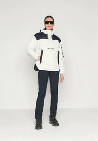 Hvitt og marineblått jakke med høy krage, frontlomme og logo. Kombinert med mørke bukser og svarte tursko. Jevn tekstur.