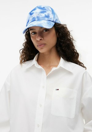 Mujer con cabello rizado que lleva una camisa blanca abotonada con un pequeño logo en el bolsillo y una gorra de béisbol azul teñida con técnica tie-dye de Tommy Jeans.