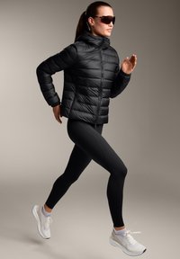 Črna pufasta jakna z horizontalnim šivanjem, eleganten dizajn in kapuco; v kombinaciji s črnimi leggins in belimi športnimi copati.