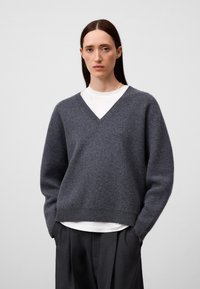 Grijze wollen V-hals trui met geribbelde zoom. Draag over een wit T-shirt. Losse pasvorm en lange mouwen. Eenvoudig ontwerp zonder versieringen.