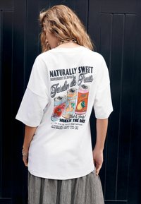 Fehér, túlméretezett póló színes italillusztrációkkal és felirattal a hátoldalán. A szövet puhanak, kényelmesnek és könnyűnek tűnik.