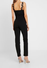 Mono negro con acentos de encaje en los tirantes, silueta ajustada, piernas estrechas y una textura suave. Llevado con tacones altos plateados con tiras.