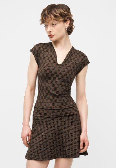 Γυναίκα που φοράει εφαρμοστό καφέ φόρεμα με μαύρα γεωμετρικά σχέδια, κοντά μανίκια και V-λαιμόκοψη, στέκεται μπροστά σε απλό ανοιχτόχρωμο φόντο.