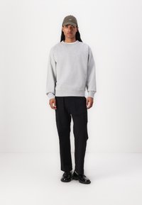 Felpa grigia con colletto a girocoso, pantaloni cargo neri e scarpe nere. Il modello indossa un cappellino verde oliva. Outfit semplice e casual.