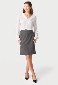 Witte blouse met verticale plissés en lange mouwen, gecombineerd met een grijze tweed rok. De rok heeft een lichte split en een strakke silhouet.