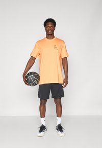 T-shirt en coton orange à manches courtes avec un petit graphique, associé à un short de sport noir. Tenant un ballon de basket, portant des chaussures noires et blanches.