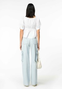 Top bianco con maniche a puff e vita arricciata; abbinato a pantaloni cargo a gamba larga blu chiaro con tasche laterali; modello che tiene in mano una piccola borsa bianca.