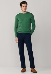 Grüner Pullover mit Rundhalsausschnitt, kombiniert mit marineblauer Hose und braunen Wildlederschuhen. Der Stoff wirkt weich und körpernah geschnitten.