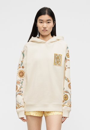 Hosszú sötét hajú nő, krémszínű kapucnis pulóverben, amely nap- és virágmintákat díszít az ujjakon és a mellkasi zsebén, sárga mintás rövidnadrággal párosítva.