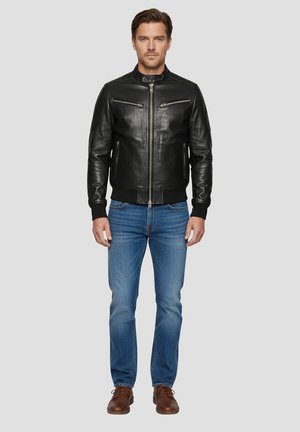 Sort læder bomberjakke med lynlås, to brystlommer, ribstrikkede manchetter og kant, kombineret med blå jeans og brune sko.