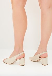 Beige avokkaat, joissa on auki oleva varvas ja kiinnitys mikrokuitumateriaalista, ja jotka ovat varustettu lohkarekorolla sekä minimalistisella muotoilulla. Säädettävä hihna näkyy takana.
