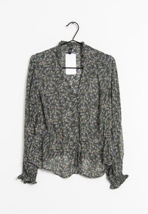 Blouse florale en tissu vert foncé, avec un ourlet noué à l'avant, des manches longues avec des poignets volants, et un motif de fleurs subtil en rose et jaune.