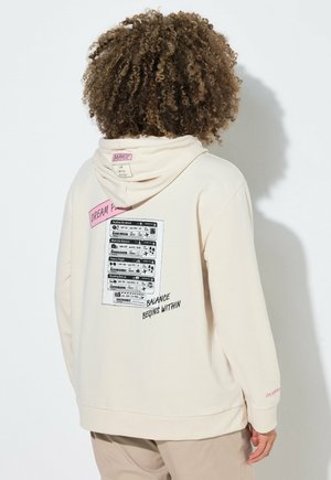 Person mit lockigem Haar trägt einen cremefarbenen Hoodie und zeigt den Rücken mit aufgedrucktem Text und Grafiken über Balance und Wohlbefinden.