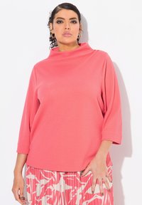 Maglione corallo con colletto alto e maniche tre quarti. Tessuto con texture e vestibilità morbida, abbinato a una gonna a fantasia.
