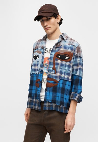 Καρό φανελένιο πουκάμισο σε μπλε και καφέ με καλλιτεχνικά σχέδια προσώπων. Διαθέτει αντίθετες υφές, ξεφτισμένες άκρες και κουμπιά μπροστά. Καφέ καπέλο.