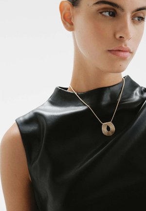 Collar de oro con un diseño ovalado texturizado, lucido por un modelo que lleva una camiseta sin mangas de cuero negro, destacando una estética moderna y elegante.
