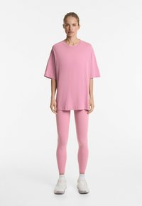 Tricou oversized roz deschis cu mâneci scurte, asortat cu colanți roz. Ambele sunt realizate dintr-un material neted și elastic. Adida�și albi incluși.