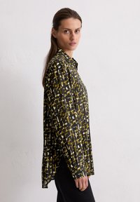 Femme aux longs cheveux bruns portant une chemise à motifs, ample et à boutons, avec des formes abstraites noires, vert olive et blanches, associée à un pantalon noir.