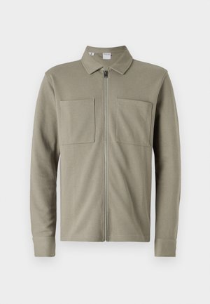 Veste zippée vert olive en tissu texturé, avec un col et deux grandes poches avant. Manches longues avec des ourlets à double retour.