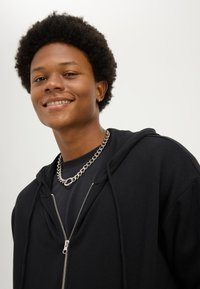 Hymyilevä nuori afrohiuksinen henkilö pukeutuu mustaan huppariin, mustaan paitaan, hopeiseen kaulaketjuun ja pieneen korvarenkaaseen yksivärisen taustan edessä.