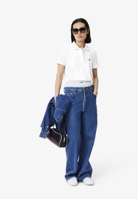Polo bianco con un piccolo logo, jeans in denim oversize blu, giacca di denim, borsa nera e sneakers bianche. Occhiali da sole scuri completano il look.