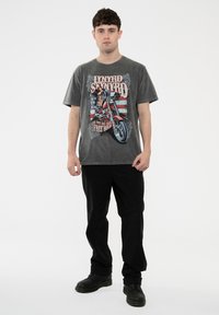 Gray cotton t-shirt featuring a motorcycle graphic and text against a faded American flag background. Black pants and dark footwear complement the look.