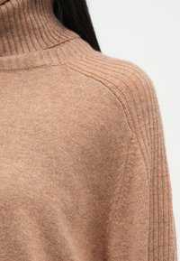 Μπλούζα σε μαλακό, ανοιχτό μπεζ ύφασμα με ραβδωτό ψηλό λαιμό. Περιλαμβάνει πτώσεις στους ώμους και λεία υφή.