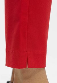 Rote Stoffhose mit strukturierter Textur und einem seitlichen Schlitz am Saum. Das Design ist maßgeschneidert und betont eine glatte, figurbetonte Silhouette.