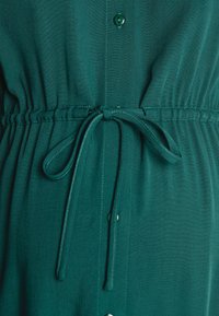 Vestido de tecido verde, leve, com botão frontal, cintura elástica com cordão ajustável e textura suave.