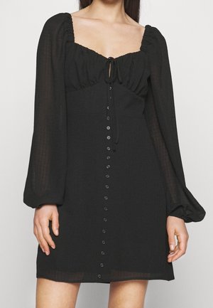 Vrouw draagt een zwarte minijurk met lange mouwen, geplooide halslijn, strik aan de voorkant en een rij knopen in het midden.