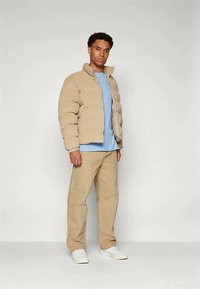 Vyras stovi vilkėdamas smėlio spalvos pūkinę striukę, šviesiai mėlyną marškinį, smėlio spalvos kelnes ir baltus sportinius batus, prieš paprastą šviesų foną.