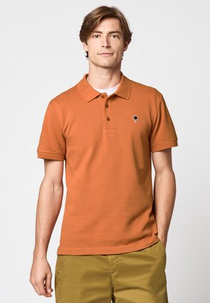 Jeune homme portant un polo à manches courtes couleur rouille avec un petit logo d'arbre noir et un pantalon jaune moutarde, debout devant un fond uni.