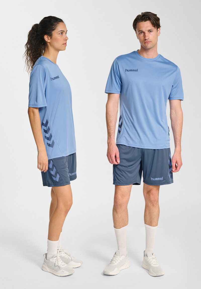 Twee modellen die bijpassende lichtblauwe sportshirts en donkerblauwe shorts dragen met witte sneakers, staand tegen een witte achtergrond.