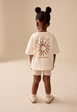Barn med to afro-puff-frisurer iført en stor, cremefarvet T-shirt med solmotiv og fransk tekst, stribede shorts og sneakers, set bagfra.