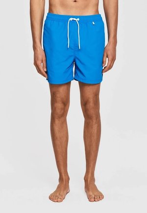 Mężczyzna ubrany w jasnoniebieskie kąpielówki z białym sznurkiem, stojący boso na jednolitym jasnym tle.