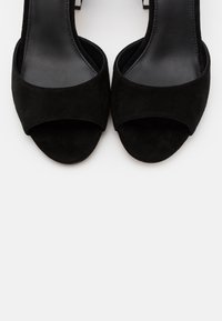Paire de sandales ouvertes en suède noir avec une large bride avant et une semelle intérieure noire lisse sur un fond clair uni.
