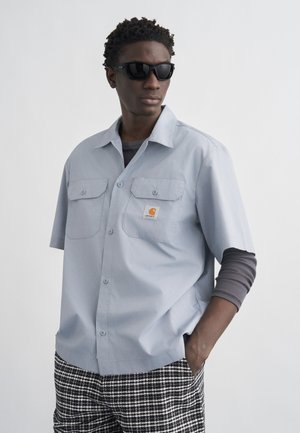 Hombre con gafas de sol negras, camisa de manga corta azul claro abotonada sobre manga larga gris, y pantalones de cuadros en blanco y negro, con la mano en el bolsillo.