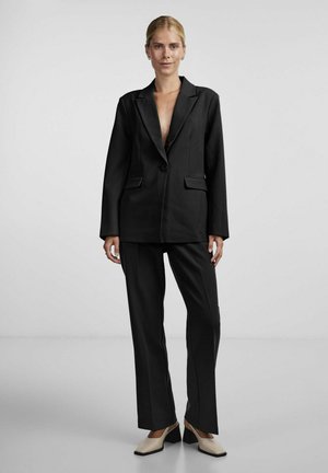 Femme portant un costume noir sur mesure avec un blazer à un bouton et un pantalon droit, associée à des chaussures beige à talons bas, debout devant un fond uni.