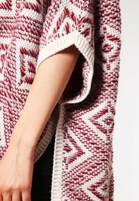 Strikket poncho med et geometrisk mønster i bordeaux og creme. Har korte ærmer og en løs pasform, fremstillet af tekstureret garn.