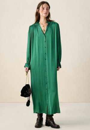 Grünes langärmeliges Kleid aus glattem Stoff mit Knopfleiste, plissierten Schultern und Manschetten. Kombiniert mit einer schwarzen Handtasche und Stiefeln.