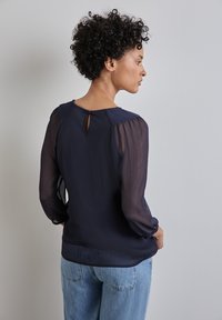 Blusa navy con maniche a sbuffo trasparenti e dettaglio a apertura a chiave sul retro. Tessuto testurizzato con vestibilità ampia, abbinata a jeans azzurri chiari.