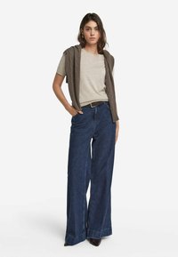 Lys beige t-shirt parret med højtaljede mørkeblå brede jeans og en brun strikket vest hængende over skuldrene.