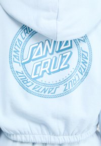 Svetlomodrá mikina s kapucňou s veľkým kruhovým logom tmavšej modrej farby, na ktorom je nápis "Santa Cruz Skateboards", s bodkovými detailmi a elastickým pásom.