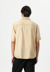 Camisa de botones de manga corta en amarillo claro, hecha de una tela suave con una textura ligeramente arrugada y un dobladillo curvado. Vista trasera visible.
