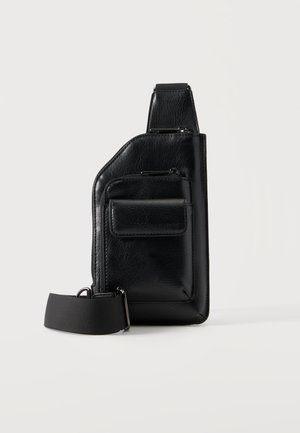Sac bandoulière en cuir noir avec deux compartiments zippés, une poche avant avec rabat et une sangle en tissu réglable sur fond blanc.