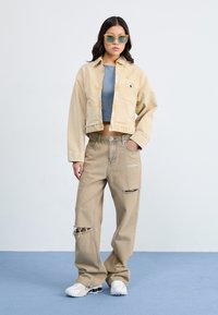 Veste en denim beige coupée avec poche avant, haut gris côtelé, pantalon large beige usé, et baskets blanches avec accents noirs.
