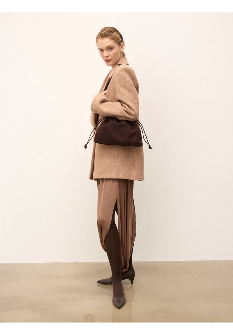 Béžový vlnený kabát cez hnedé drapované nohavice. Hnedá taška na šnúrku a tlmené hnedé lodičky. Hladké textúry a neutrálna farebná paleta.